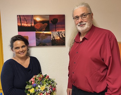 Ein Bild, welches einen Mann rechts und eine Frau links zeigt. Die beiden stehen vor einer Wand, an der ein farbenfrohes Bild hngt. Die Frau hlt einen Blumenstrau in den Hnden, und beide lcheln und schauen sehr freundlich in die Kamera.